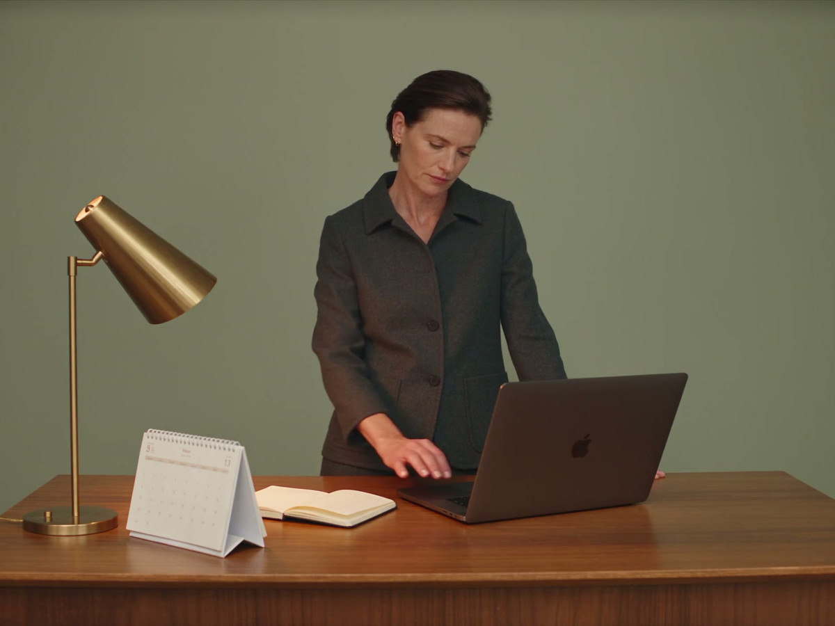 A woman checks a desk calendar and uses the trackpad on her laptop. The screen faces her.