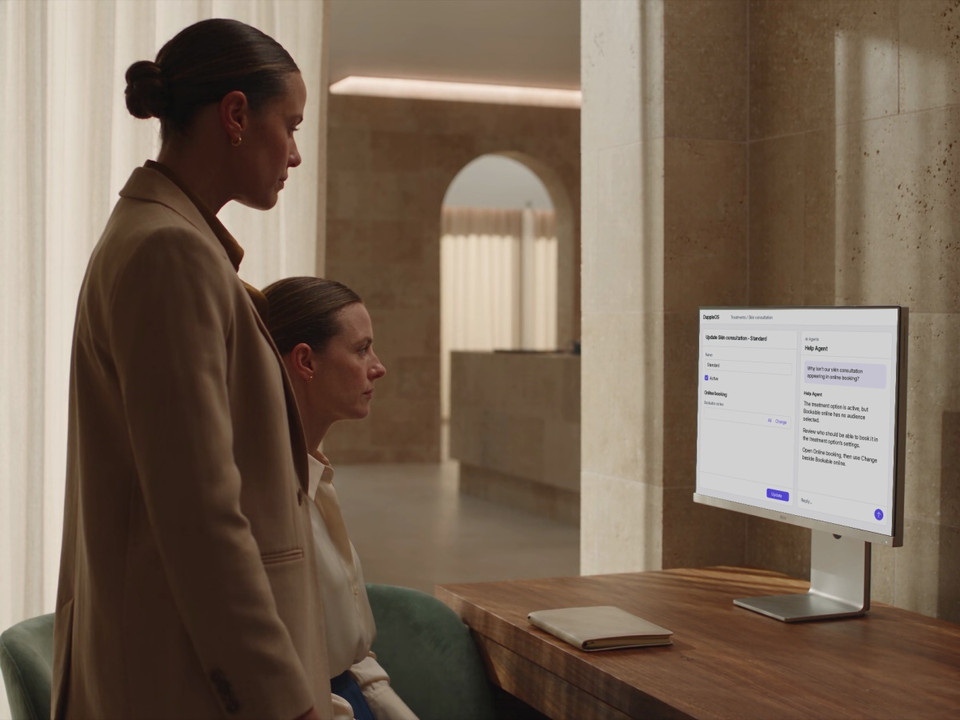 Two clinic colleagues review an unchanged online-booking setting beside Help Agent guidance on a desktop monitor.