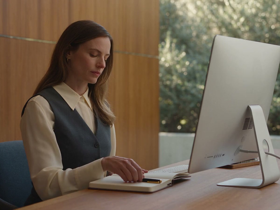 A clinic manager reviews the day at a supported desktop computer and records a question in her notebook
